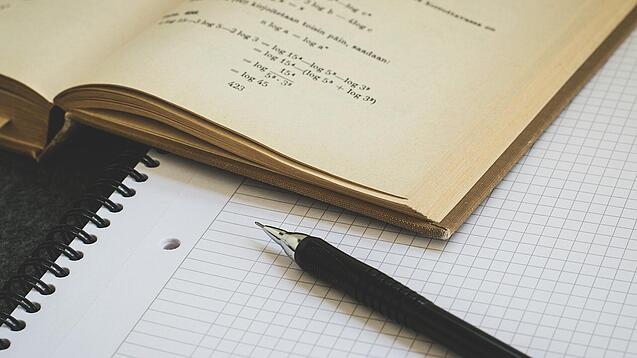 Ein Buch liegt aufgeschlagen auf einem Notizblock, auf dem auch ein Stift liegt.
