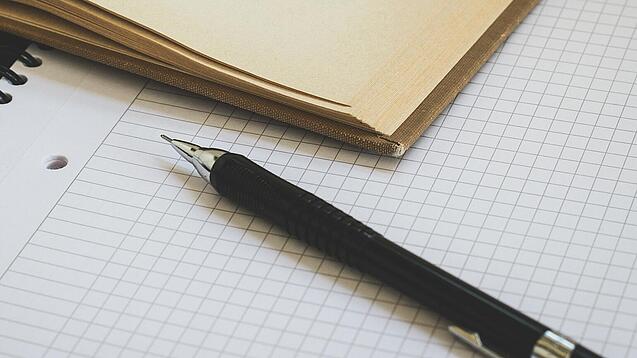 Ein Buch liegt aufgeschlagen auf einem Notizblock, auf dem auch ein Stift liegt.