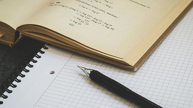 Ein Buch liegt aufgeschlagen auf einem Notizblock, auf dem auch ein Stift liegt.