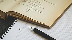 Ein Buch liegt aufgeschlagen auf einem Notizblock, auf dem auch ein Stift liegt.