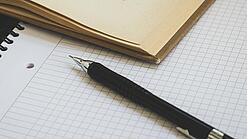 Ein Buch liegt aufgeschlagen auf einem Notizblock, auf dem auch ein Stift liegt.