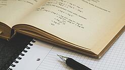 Ein Buch liegt aufgeschlagen auf einem Notizblock, auf dem auch ein Stift liegt.