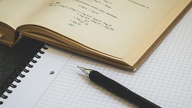 Ein Buch liegt aufgeschlagen auf einem Notizblock, auf dem auch ein Stift liegt.