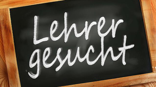 Auf einer kleinen Schiefertafel mit braunem Holzrand steht mit weißer Kreide "Lehrer gesucht" geschrieben.