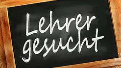 Auf einer kleinen Schiefertafel mit braunem Holzrand steht mit weißer Kreide "Lehrer gesucht" geschrieben.