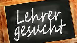 Auf einer kleinen Schiefertafel mit braunem Holzrand steht mit weißer Kreide "Lehrer gesucht" geschrieben.