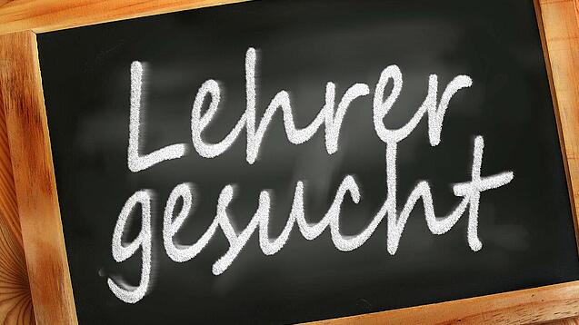 Auf einer kleinen Schiefertafel mit braunem Holzrand steht mit weißer Kreide "Lehrer gesucht" geschrieben.