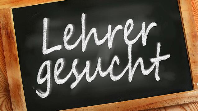 Auf einer kleinen Schiefertafel mit braunem Holzrand steht mit weißer Kreide "Lehrer gesucht" geschrieben.
