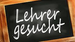 Auf einer kleinen Schiefertafel mit braunem Holzrand steht mit weißer Kreide "Lehrer gesucht" geschrieben.