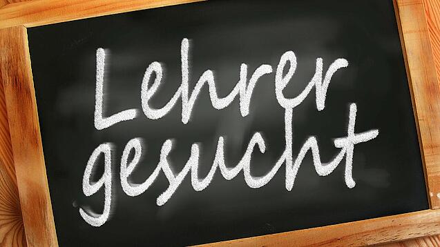 Auf einer kleinen Schiefertafel mit braunem Holzrand steht mit weißer Kreide "Lehrer gesucht" geschrieben.