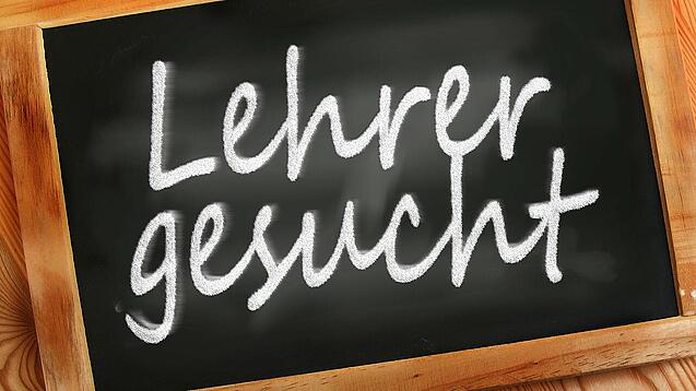 Auf einer kleinen Schiefertafel mit braunem Holzrand steht mit weißer Kreide "Lehrer gesucht" geschrieben.