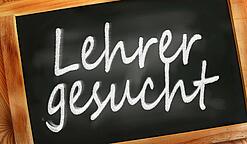 Auf einer kleinen Schiefertafel mit braunem Holzrand steht mit weißer Kreide "Lehrer gesucht" geschrieben.