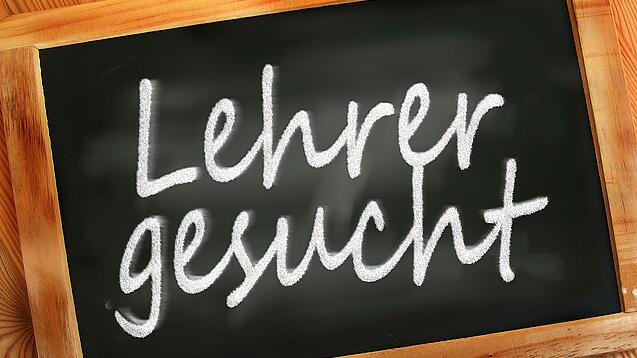 Auf einer kleinen Schiefertafel mit braunem Holzrand steht mit weißer Kreide "Lehrer gesucht" geschrieben.