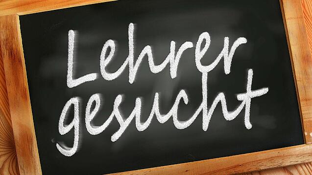 Auf einer kleinen Schiefertafel mit braunem Holzrand steht mit weißer Kreide "Lehrer gesucht" geschrieben.