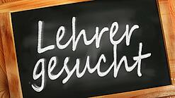 Auf einer kleinen Schiefertafel mit braunem Holzrand steht mit weißer Kreide "Lehrer gesucht" geschrieben.
