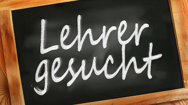 Auf einer kleinen Schiefertafel mit braunem Holzrand steht mit weißer Kreide "Lehrer gesucht" geschrieben.