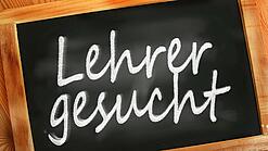 Auf einer kleinen Schiefertafel mit braunem Holzrand steht mit weißer Kreide "Lehrer gesucht" geschrieben.
