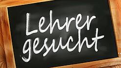 Auf einer kleinen Schiefertafel mit braunem Holzrand steht mit weißer Kreide "Lehrer gesucht" geschrieben.