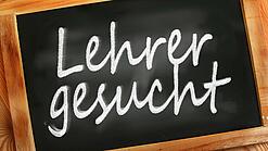 Auf einer kleinen Schiefertafel mit braunem Holzrand steht mit weißer Kreide "Lehrer gesucht" geschrieben.