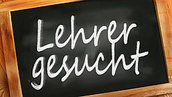 Auf einer kleinen Schiefertafel mit braunem Holzrand steht mit weißer Kreide "Lehrer gesucht" geschrieben.