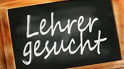 Auf einer kleinen Schiefertafel mit braunem Holzrand steht mit weißer Kreide "Lehrer gesucht" geschrieben.