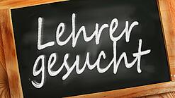 Auf einer kleinen Schiefertafel mit braunem Holzrand steht mit weißer Kreide "Lehrer gesucht" geschrieben.