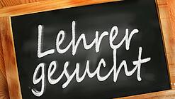Auf einer kleinen Schiefertafel mit braunem Holzrand steht mit weißer Kreide "Lehrer gesucht" geschrieben.