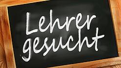 Auf einer kleinen Schiefertafel mit braunem Holzrand steht mit weißer Kreide "Lehrer gesucht" geschrieben.