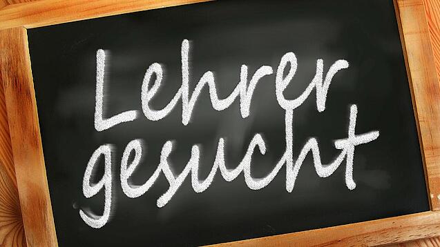 Auf einer kleinen Schiefertafel mit braunem Holzrand steht mit weißer Kreide "Lehrer gesucht" geschrieben.