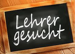 Auf einer kleinen Schiefertafel mit braunem Holzrand steht mit weißer Kreide "Lehrer gesucht" geschrieben.