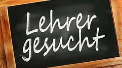 Auf einer kleinen Schiefertafel mit braunem Holzrand steht mit weißer Kreide "Lehrer gesucht" geschrieben.