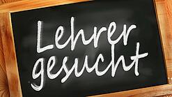 Auf einer kleinen Schiefertafel mit braunem Holzrand steht mit weißer Kreide "Lehrer gesucht" geschrieben.