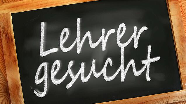 Auf einer kleinen Schiefertafel mit braunem Holzrand steht mit weißer Kreide "Lehrer gesucht" geschrieben.