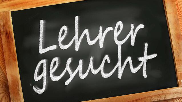 Auf einer kleinen Schiefertafel mit braunem Holzrand steht mit weißer Kreide "Lehrer gesucht" geschrieben.