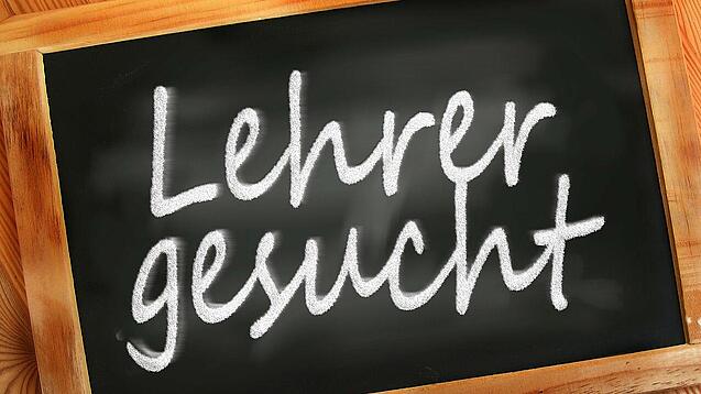 Auf einer kleinen Schiefertafel mit braunem Holzrand steht mit weißer Kreide "Lehrer gesucht" geschrieben.