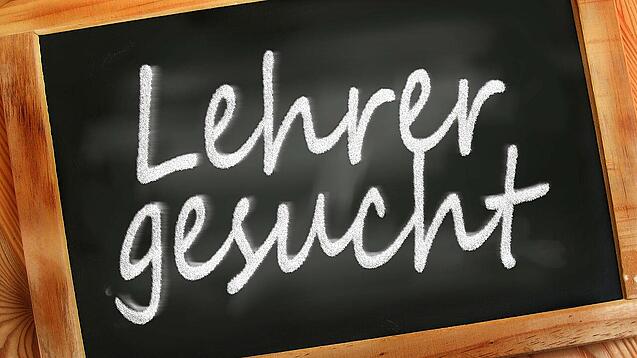 Auf einer kleinen Schiefertafel mit braunem Holzrand steht mit weißer Kreide "Lehrer gesucht" geschrieben.