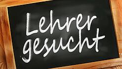 Auf einer kleinen Schiefertafel mit braunem Holzrand steht mit weißer Kreide "Lehrer gesucht" geschrieben.