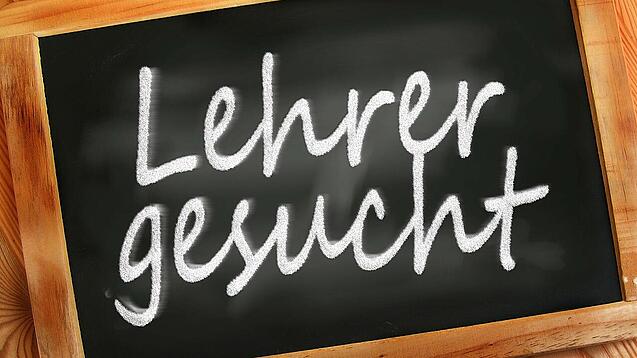 Auf einer kleinen Schiefertafel mit braunem Holzrand steht mit weißer Kreide "Lehrer gesucht" geschrieben.