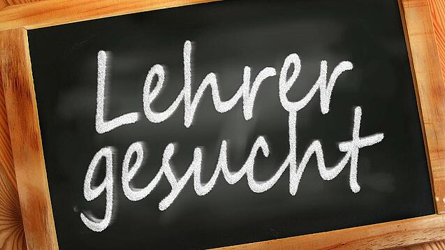 Auf einer kleinen Schiefertafel mit braunem Holzrand steht mit weißer Kreide "Lehrer gesucht" geschrieben.