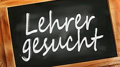 Auf einer kleinen Schiefertafel mit braunem Holzrand steht mit weißer Kreide "Lehrer gesucht" geschrieben.