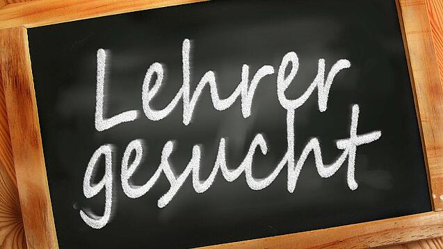 Auf einer kleinen Schiefertafel mit braunem Holzrand steht mit weißer Kreide "Lehrer gesucht" geschrieben.