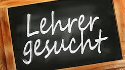 Auf einer kleinen Schiefertafel mit braunem Holzrand steht mit weißer Kreide "Lehrer gesucht" geschrieben.