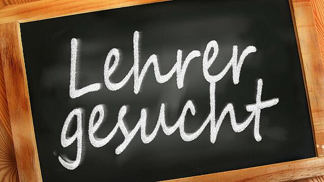 Auf einer kleinen Schiefertafel mit braunem Holzrand steht mit weißer Kreide "Lehrer gesucht" geschrieben.