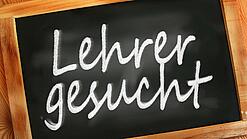 Auf einer kleinen Schiefertafel mit braunem Holzrand steht mit weißer Kreide "Lehrer gesucht" geschrieben.