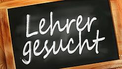 Auf einer kleinen Schiefertafel mit braunem Holzrand steht mit weißer Kreide "Lehrer gesucht" geschrieben.