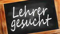 Auf einer kleinen Schiefertafel mit braunem Holzrand steht mit weißer Kreide "Lehrer gesucht" geschrieben.