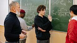 Studierende und ein Dozent lösen Mathematikaufgaben an einer Tafel.