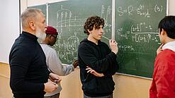 Studierende und ein Dozent lösen Mathematikaufgaben an einer Tafel.